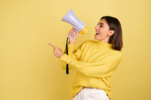 Dizione.it Celebra La Giornata Mondiale Della Voce 9 Giovane Donna In Maglione Giallo Caldo Con Altoparlante Megafono Che Urla A Sinistra Puntando Il Dito Indice 1