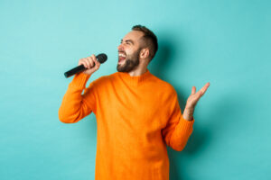 Il Training Vocale: Cosa Significa Allenare La Voce 2 Bell Uomo Adulto Esegue Una Canzone Cantando Nel Microfono In Piedi Contro Il Muro Turchese 1